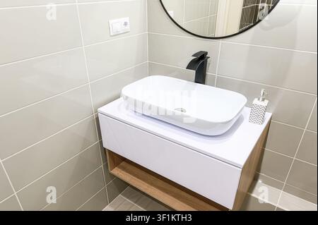 modern bathroom features a white rectangular sink, black faucet, and gray tiles. A round mirror hangs above Stock Photo