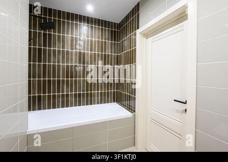 modern bathroom with a white bathtub, dark vertical tiles in the shower area, and light grey tiles on the walls. A white door with a black handle is v Stock Photo