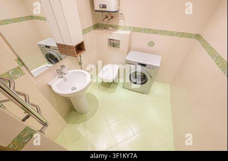 compact bathroom with pastel tiles, featuring a pedestal sink, toilet, and washing machine. A narrow mirror hangs above the sink, reflecting the laund Stock Photo