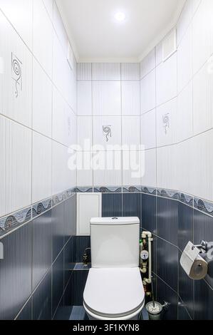 small tiled bathroom featuring a white toilet, with blue and white tiles. The top of the walls are white with unique drawings, and the bottom of the w Stock Photo