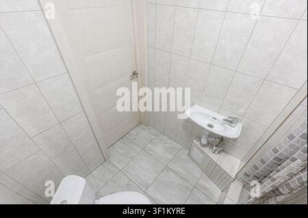 clean, tiled bathroom features a toilet, sink, and door. The space is brightly lit and has a minimalist design with light grey and white tones Stock Photo
