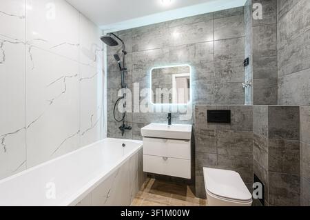 modern bathroom with white tub, sink, toilet, and a lighted mirror. Walls are tiled in white marble and gray stone Stock Photo