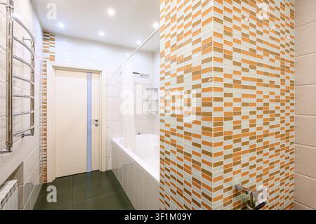 well-lit bathroom features a tub with a shower curtain, a heated towel rack, and a striking mosaic tiled wall in shades of orange, beige, and white Stock Photo