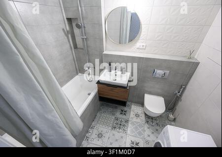 Modern bathroom with grey and white tiles, patterned floor, bathtub with shower, round mirror, and floating vanity. Clean, contemporary design Stock Photo