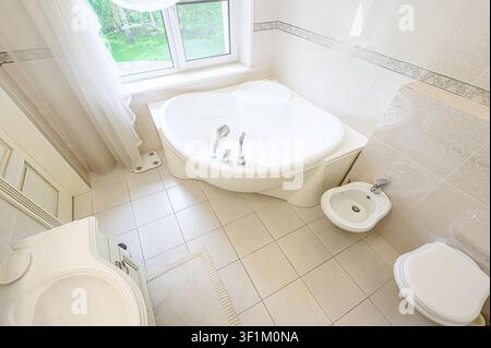 Bright bathroom with a large white tub, bidet, toilet, sink, and window. Tiled floor and walls Stock Photo