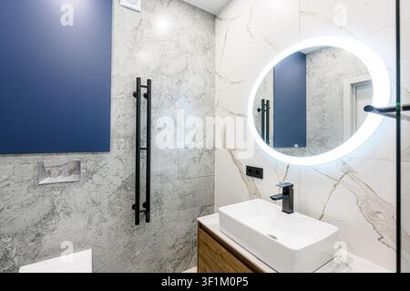 Modern bathroom with marble tiles, a white rectangular sink, black faucet, round LED mirror, and a blue accent panel Stock Photo