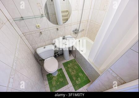 bathroom features a toilet, sink, bathtub, a shower curtain, and light-colored tiles. A mirror and green mats are also visible Stock Photo