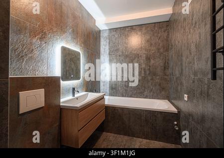 modern bathroom featuring textured, earth-toned tiles, a sleek sink with a wooden cabinet, an illuminated mirror, and a white bathtub Stock Photo
