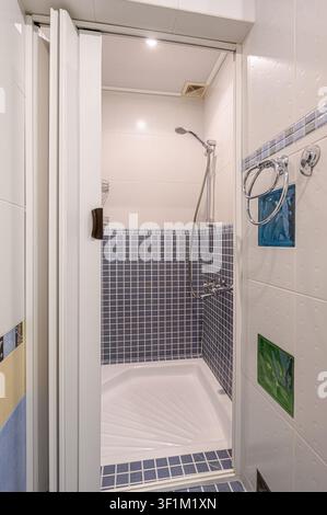 compact shower stall with blue-tiled walls and a white shower base. A white door is partially open, revealing a chrome showerhead Stock Photo