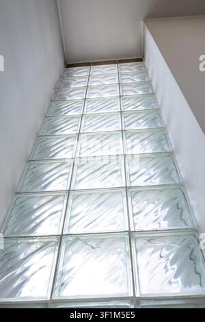 A low-angle, sharp perspective shot of a tall vertical glass block window wall framed by white plaster walls, showing bright diffused daylight shining Stock Photo