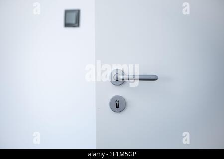 A close-up shot of a modern matte silver door handle and keyhole on a minimalist white door slightly open, with a blurry light switch on the wall in t Stock Photo
