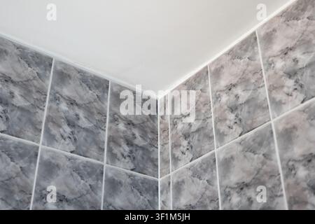 A close-up, low-angle shot focusing on the upper corner of a modern bathroom, where square grey marble-effect ceramic wall tiles meet a clean, plain w Stock Photo