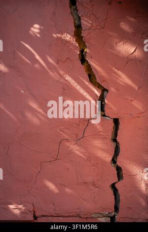 Vertical close up of a large deep crack in a pink plastered wall with leaf shadows Stock Photo
