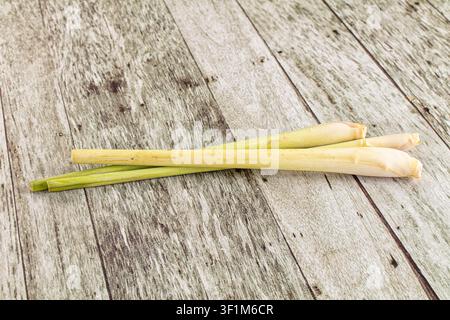Three fresh lemongrass stalks arranged diagonally on rustic wooden planks. Natural lighting highlights pale yellow green hues. Minimalist composition Stock Photo