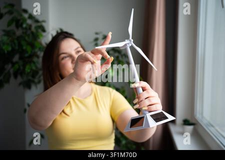 Young woman holding a small wind turbine model indoors near a window. Renewable energy education and green technology concept. Stock Photo
