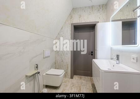 Bright bathroom featuring a toilet, sink, illuminated mirror, and a gray door, with mixed tile patterns on the walls and floor Stock Photo