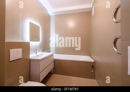 bathroom features beige tiles, a white sink vanity with an illuminated mirror, and a bathtub. Modern, clean design Stock Photo