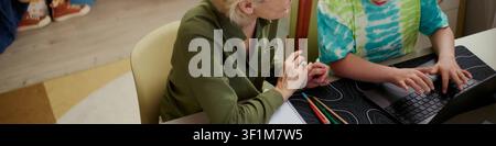 Senior Caucasian woman guiding child using laptop at desk, supporting digital learning and classroom mentoring. Useful for education technology, tutoring, parenting campaigns Stock Photo