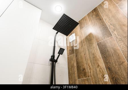 low-angle shot of a modern shower with black fixtures, contrasting wood-look tiles, and white tiles. A ceiling light illuminates the scene Stock Photo