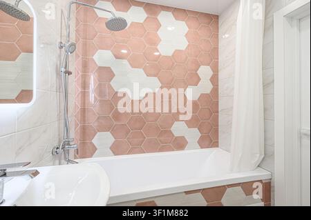 modern bathroom features pink and white hexagon tiles, a white bathtub, a sink with a mirror, and a showerhead Stock Photo