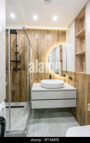 Modern bathroom with wood-look tile, round lighted mirror, white vanity, shower, and marble floor Stock Photo