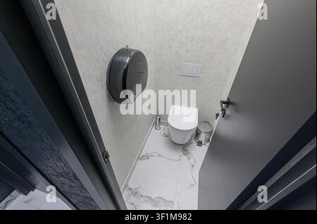 modern toilet features a white toilet, marble tile floor, gray walls with dispenser, and a dark door ajar Stock Photo