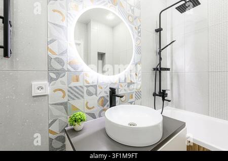 Modern bathroom with white suite, round lighted mirror, black fixtures, and stylish patterned & textured tiles Stock Photo