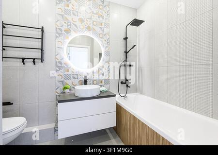 Modern bathroom with white suite, round lighted mirror, black fixtures, and stylish patterned & textured tiles Stock Photo
