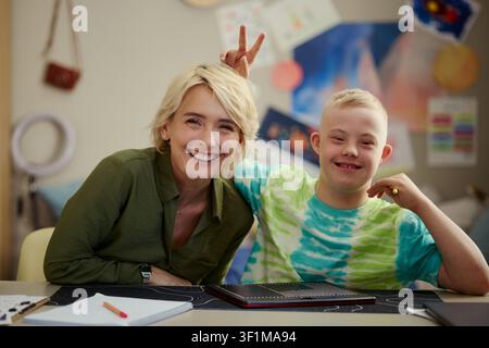 Portrait of Caucasian teacher and child with Down Syndrome smiling at camera, sitting at desk during classroom activity. Useful for inclusive education, support service, caregiving Stock Photo