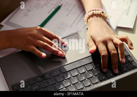 Close up of child hands using laptop touchpad and keyboard beside worksheet, practicing digital learning and homework. Suitable for education technology, online study, parenting campaigns Stock Photo