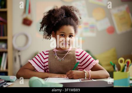 Portrait of Black girl studying and writing at desk, looking at camera while doing schoolwork. Ideal for education, childhood learning, tutoring, classroom promotion campaigns Stock Photo