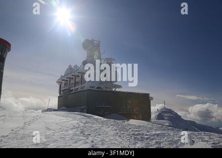 Bright sunburst shines over a heavily iced telecommunications tower and station building at the summit of a snowy mountain peak in the Low Tatras, Slo Stock Photo