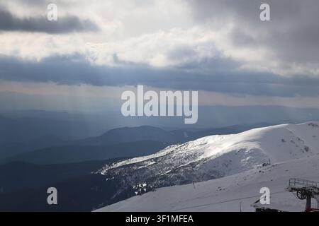 Sunbeams break through dramatic clouds illuminating a snow-covered ridge and distant mountain ranges seen from the Chopok summit area, Low Tatras, Slo Stock Photo