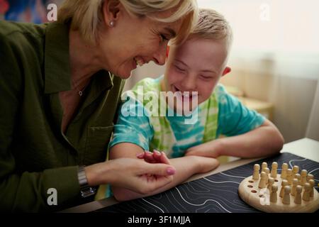 Middle aged Caucasian woman guiding child with Down Syndrome during tabletop game, supporting learning and bonding. Useful for inclusive education, caregiving, family support marketing Stock Photo