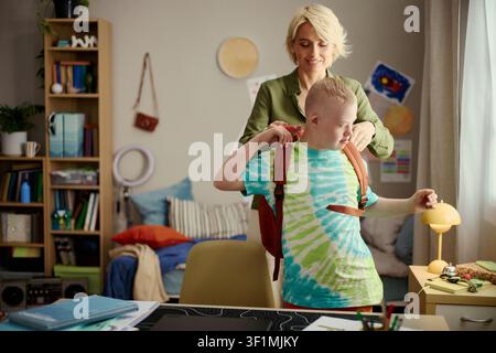 Caucasian mother helping child with Down Syndrome putting on backpack at home, supporting school readiness and independence. Useful for parenting, inclusion, education campaigns Stock Photo