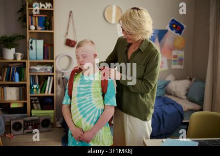Caucasian mother helping child with Down Syndrome adjusting backpack before school at home, supporting daily routine and independence. Ideal for inclusive parenting or education campaigns Stock Photo