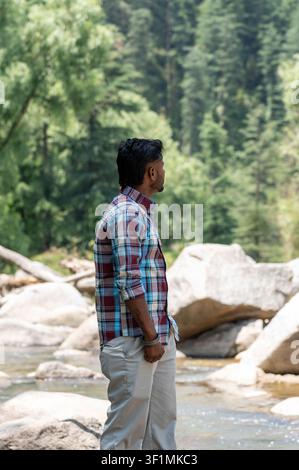 A vertical, three-quarter shot of a young Indian man wearing a maroon and blue plaid shirt, captured while looking into the distance while standing. Stock Photo