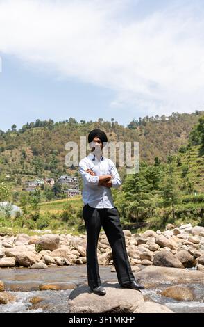 A vertical, full-length shot of a young Indian Sikh man in formal clothing standing with his arms crossed on a rock in a mountain stream. Stock Photo