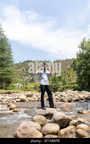 Vertical view of a young Indian Sikh man wearing formal clothing standing on rocks in a mountain stream, twirling his mustache. Stock Photo