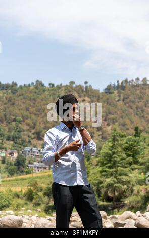 A vertical, three-quarter shot of a young Indian Sikh man in formal clothing twirling his mustache while looking away, standing outdoors. Stock Photo