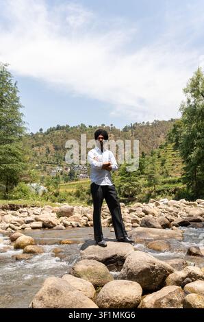 A vertical, full-length shot of a young Indian Sikh man in formal clothing standing with his arms crossed while looking at the camera. Stock Photo