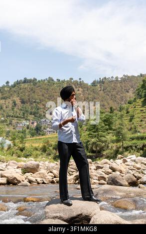 A vertical, full-length shot of a young Indian Sikh man in formal clothing twirling his mustache while looking away, standing outdoors. Stock Photo