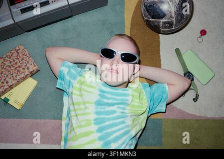 Portrait of Caucasian boy lying on floor wearing sunglasses, relaxing among personal items and gadgets, suggesting carefree childhood lifestyle. Top view, fitting youth marketing Stock Photo