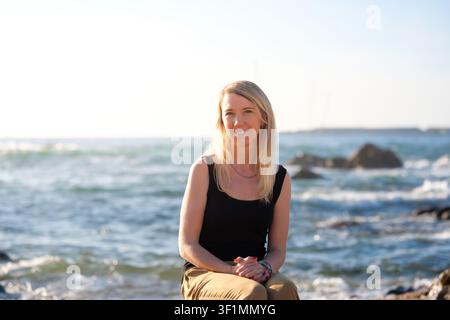 Woman sitting on rocks, enjoying sunlight and looking happily at the camera Stock Photo