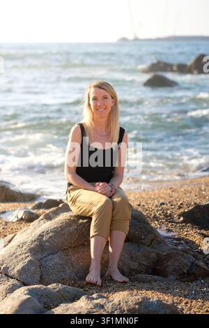 Woman smiling, sitting on a sunlit rock on the beach with ocean waves in background Stock Photo