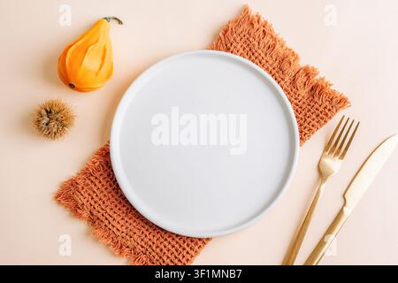 Autumn table setting mockup with empty white plate, golden cutlery, knitted napkin, decorative gourd, chestnut and copy space. Top view, flat lay. Stock Photo