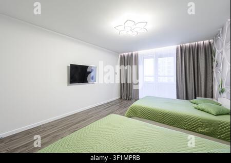 bright bedroom features two beds with light green quilted covers. A TV hangs on the white wall. Gray curtains frame a bright window Stock Photo