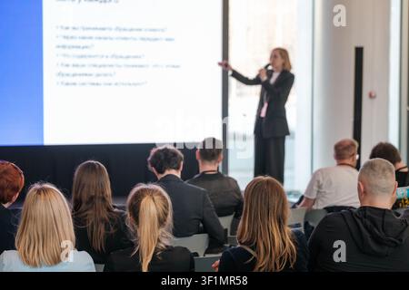 Female speaker on stage delivering business presentation to focused audience in modern conference room. Professional seminar, networking, training atmosphere. Stock Photo