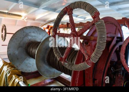 Detailed close-up of a red ship winch hand wheel wrapped with rope, beside a large steel cable spool on a working vessel deck, showing maritime indust Stock Photo