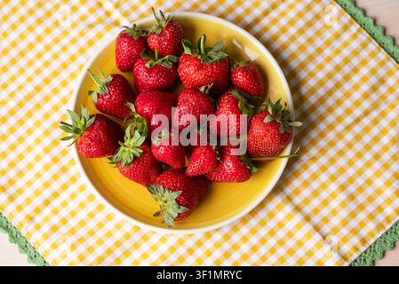 Top view of fresh strawberries with green leaves arranged in a bright yellow plate on a yellow-and-white checkered tablecloth, vibrant summer fruit sn Stock Photo